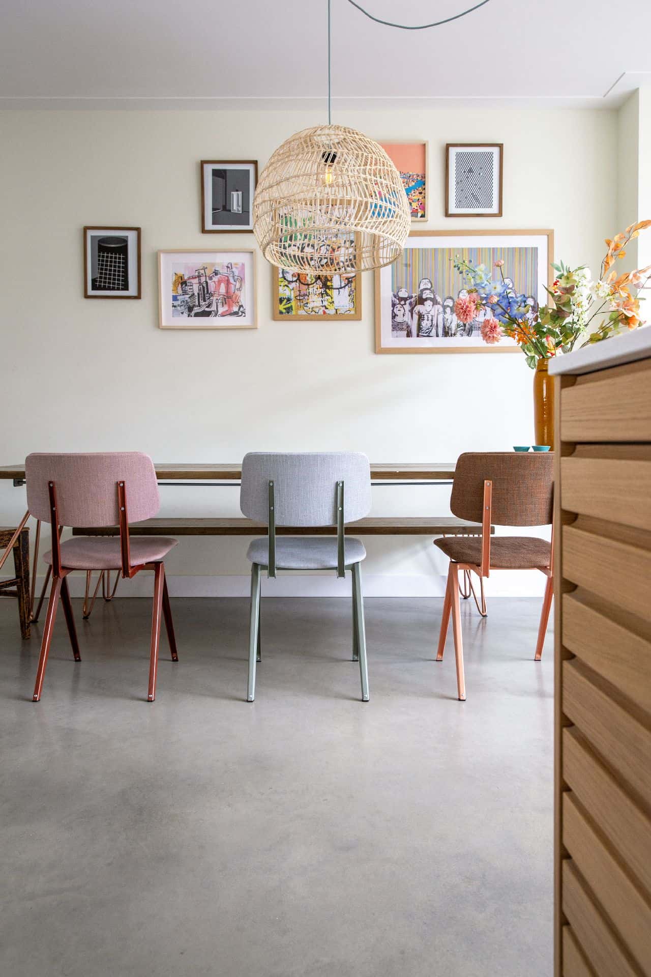 Eetkamer in Haarlem met een houten eettafel en daaraan pastel kleurige stoelen. Dit staat op een gevlinderde betonvloer. Aan de wand hangen gekleurde posters.