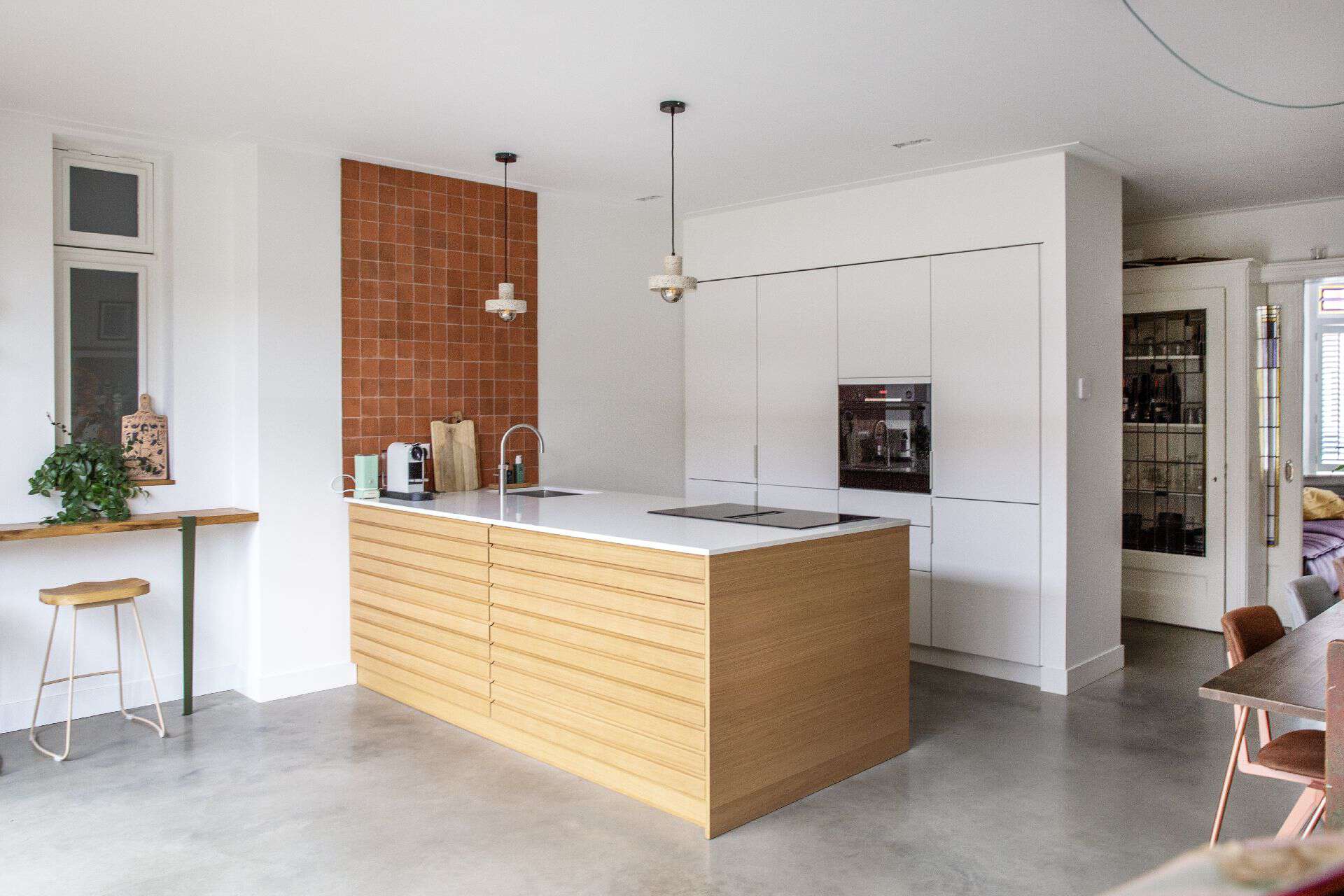 Houten kookeiland met daarachter een witte keuken in Haarlem. Tegen de witte wand zit ook een strook met oranje tegeltjes en dit is gecombineerd met een gevlinderde betonvloer