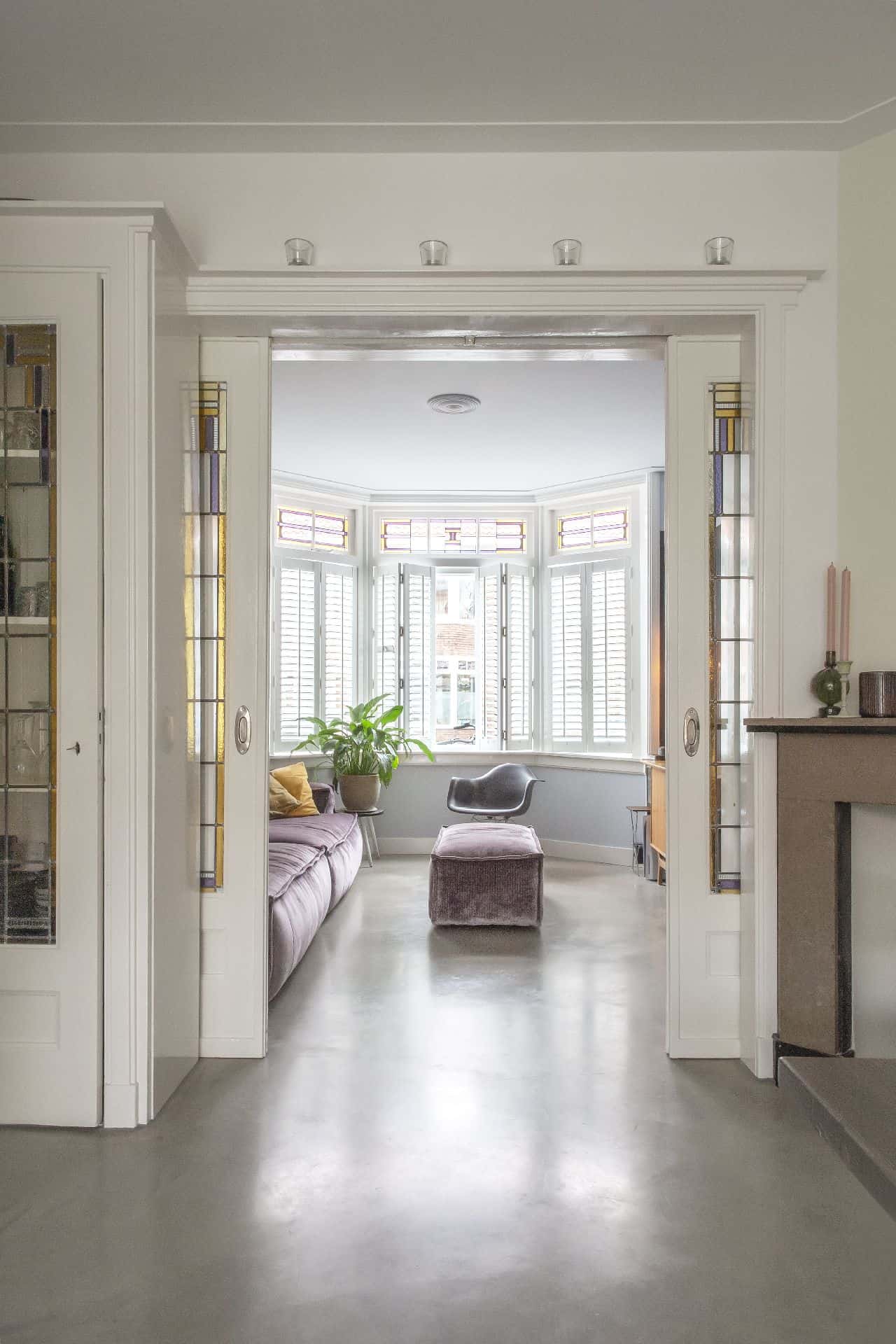 Doorkijkje naar de woonkamer. Voor de woonkamer zitten twee schuifdeuren. In de deuren zit glas in lood. Dit is gecombineerd met een gevlinderde betonvloer.