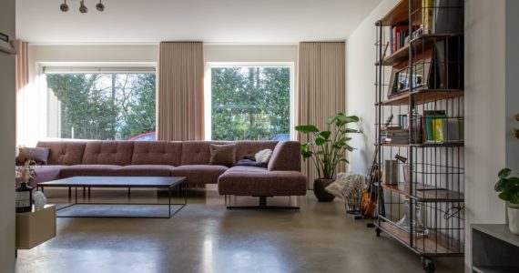 Woonkamer met een gevlinderde betonvloer in Sprang-Capelle. Ook staat er een plant op de vloer en een kast met wat boeken en accessoires.