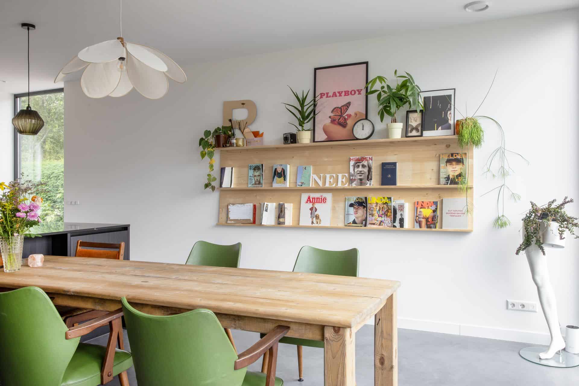 Eetgedeelte met een houten tafel en groene stoelen in Zijdewind. Achter de tafel hangt een kastje met daarin posters, platen, planten en andere accessoires. Er is gekozen voor een gevlinderde betonvloer.