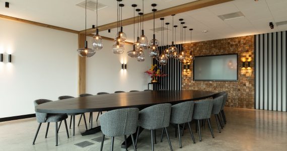 Vergaderruimte in Hattemerbroek met een gevlinderde betonvloer. Hierop staat een grote zwarte tafel met daaraan 16 grijze stoelen. Aan het plafond hangen zwarte lampjes.