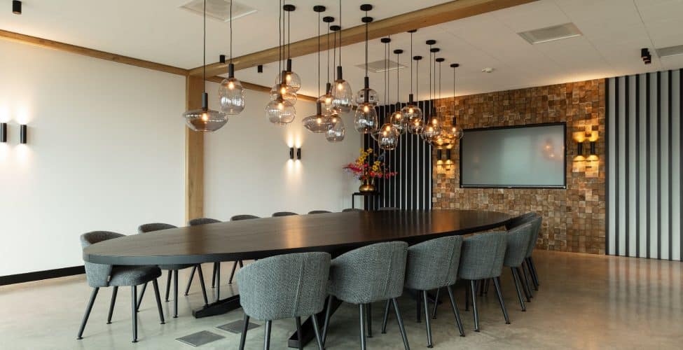 Vergaderruimte in Hattemerbroek met een gevlinderde betonvloer. Hierop staat een grote zwarte tafel met daaraan 16 grijze stoelen. Aan het plafond hangen zwarte lampjes.