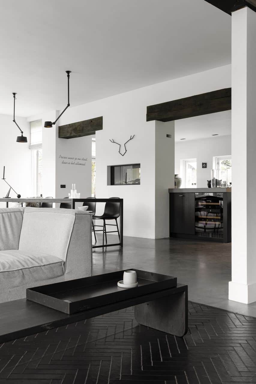 Doorkijkje van de woonkamer naar de eetkamer en de keuken. In de woonkamer staat een zwart bijzettafeltje en een grijze bank. In de eetkamer staat een zwart metalen tafel met zwarte stoelen en de keuken is zwart. Dit is gecombineerd met een gevlinderde betonvloer.
