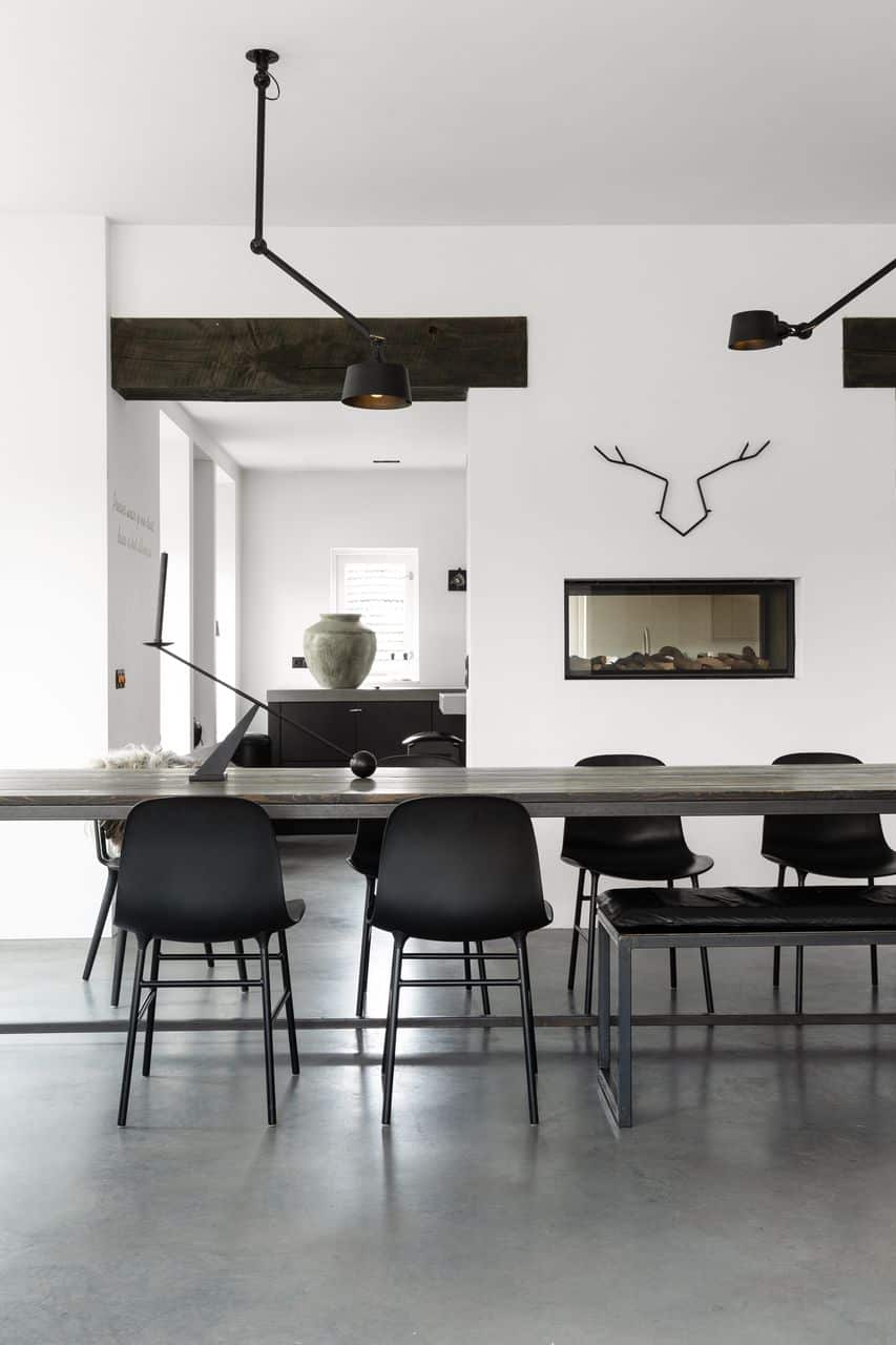 Houten tafel met een stalen frame, gecombineerd met zwarte stoelen en een gevlinderde betonvloer. De wanden zijn wit en in 1 wand zit een haard met hierboven een zwart metalen hert.