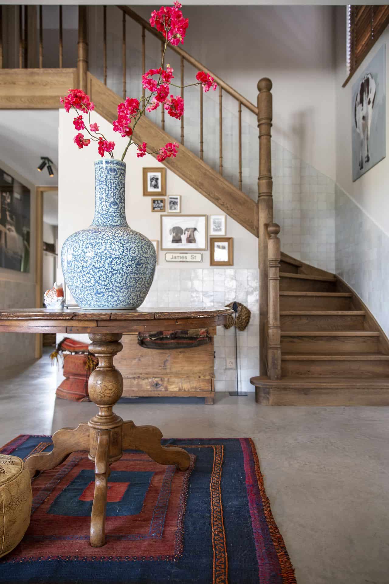 Woning in Ulvenhout met een gevlinderde betonvloer en veel houten accessoires. In deze ruimte zie je een houten tafeltje met hieronder een gekleurd vloerkleed, de houten trap en de lichte wanden.