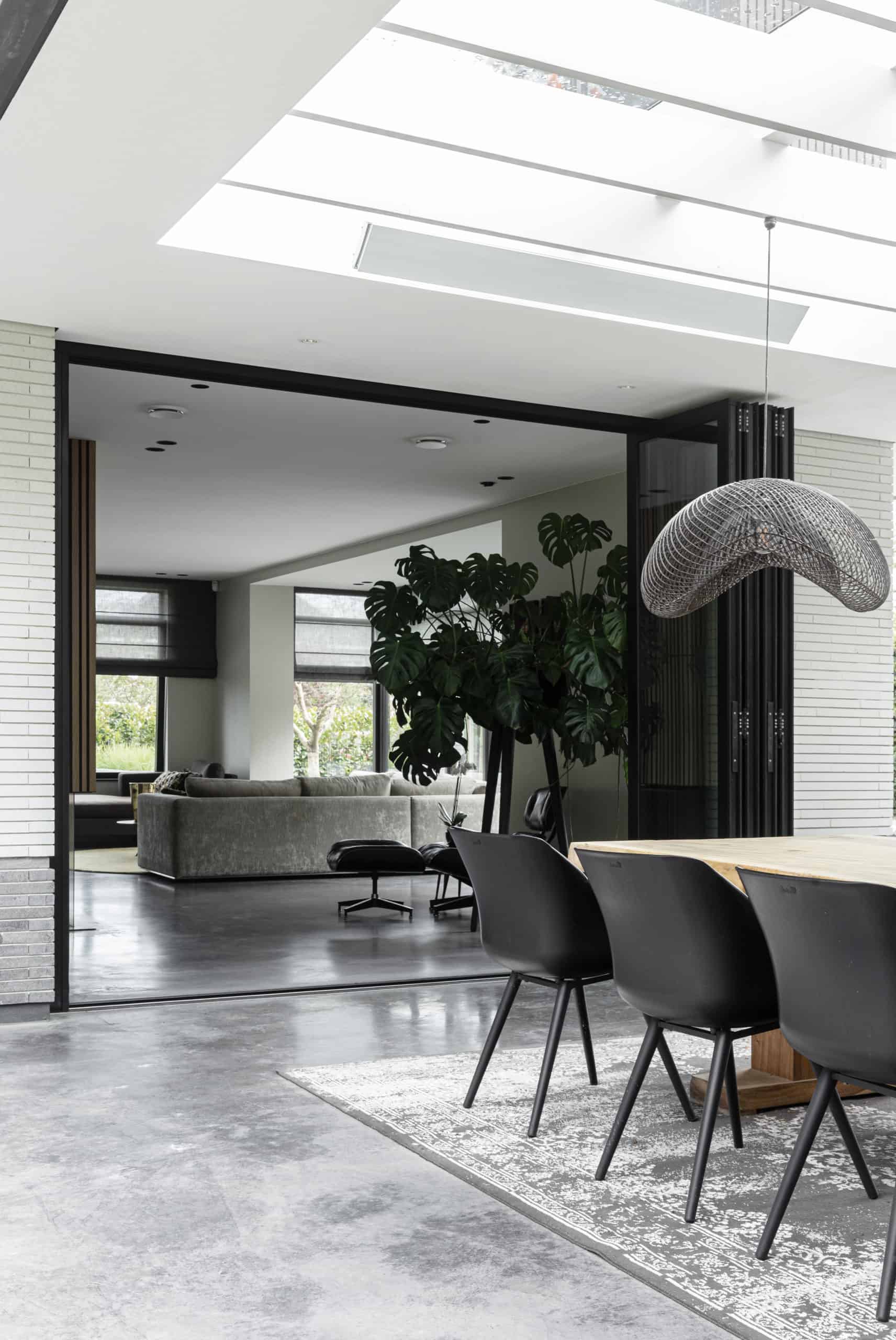 Gevlinderd betonnen terras, en gevlinderde betonvloer van Willem Designvloeren in Heemstede. Hierop staat binnen een grijze bank, en buiten ligt een vloerkleed met daarop een houten tafel en zwarte stoelen.