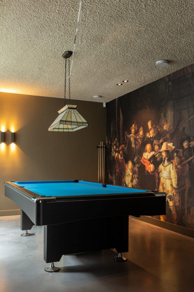 Ruimte in een villa in Heemstede met een gevlinderde betonvloer. Hierop staat een pooltafel en de wand is een grote sticker van de Nachtwacht.