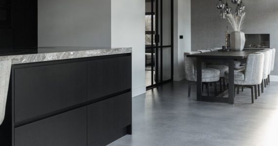 Grijze Italiaanse betonlook vloer in de kleur Rome gelegd in de landelijke keuken in Opheusden. Hierop staat een zwart kookeiland, met een grijs marmeren blad. En een zwarte tafel met daaraan grijze stoelen.