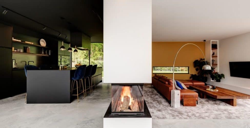 Gevlinderde betonvloer van Willem Designvloeren in de keuken en de woonkamer. Dit is gescheiden door een haard. De keuken is zwart, net als het kookeiland. Aan het kookeiland staat blauwe barkrukken. In de woonkamer ligt een groot vloerkleed met daarop een bruine bank.