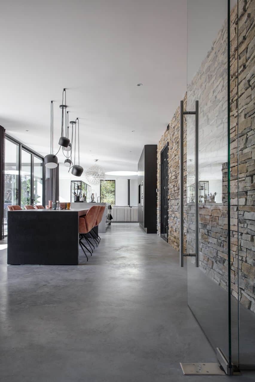 Woning in Nijmegen met een gevlinderde betonvloer in de keuken, woonkamer en eetkamer. De wand is van robuuste stenen. De eettafel is zwart met daaraan oranje stoelen.