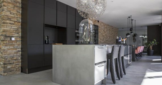 Gevlinderde betonvloer van Willem Designvloeren in Nijmegen met daarop een RVS kookeiland en een zwarte keuken in een bakstenen muur. Aan het kookeiland staat zwarte barkrukken.