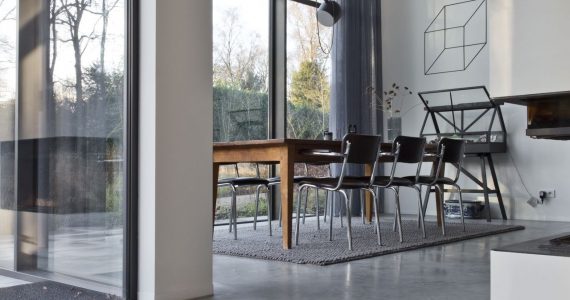 Gevlinderde betonvloer in Huizen met grote raampartijen in de eetkamer. Hier ligt een grijs vloerkleed met daarop een houten tafel en zwarte stoelen.
