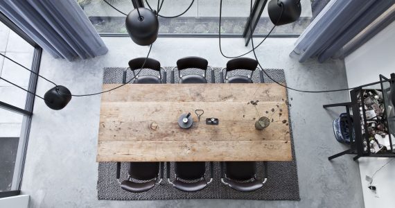 Afbeelding van bovenaf van de eetkamer in Huizen. Hier ligt een gevlinderde betonvloer met daarop een grijs vloerkleed, houten tafel en zwarte stoelen.