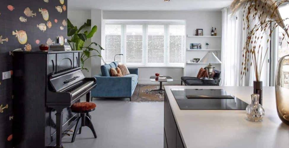Beton ciré vloer van Willem Designvloeren in de keuken en woonkamer in Kesteren. De vloer is gecombineerd met een blauwe bank en er staat een piano op de vloer.