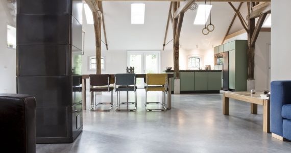 Woon boerderij in Rijswijk met een gevlinderde betonvloer van Willem Designvloeren. Hierop staat een groene keuken en een houten tafel met daaraan verschillende kleuren stoelen.