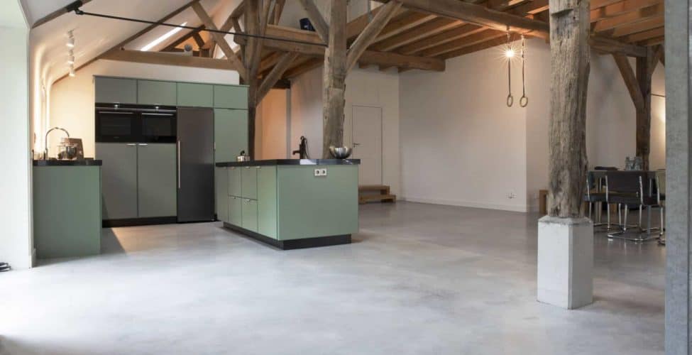 Gevlinderd betonvloer in boerderij in de keuken in Rijswijk. Hierop staat een groene keuken. Aan het plafond zitten houten balken.