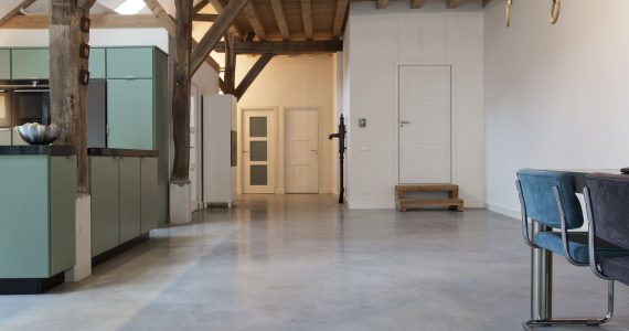 Woonboerderij in Rijswijk met een gevlinderde betonvloer van Willem Designvloeren gecombineerd met een turquoise keuken en gekleurde eetkamer stoelen.