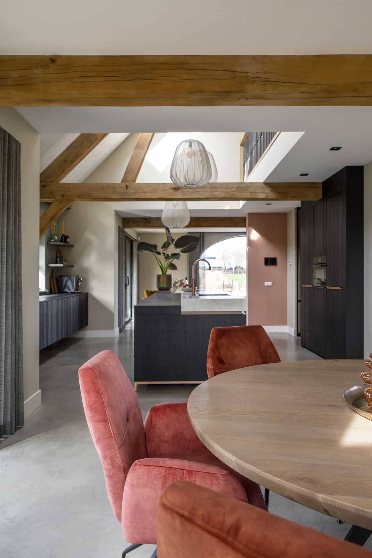 Nieuw gebouwde boerderij in Horst met een gevlinderde betonvloer. In combinatie met een zwarte keuken, houten balken aan het plafond, een houten tafel met daarom roze stoelen.
